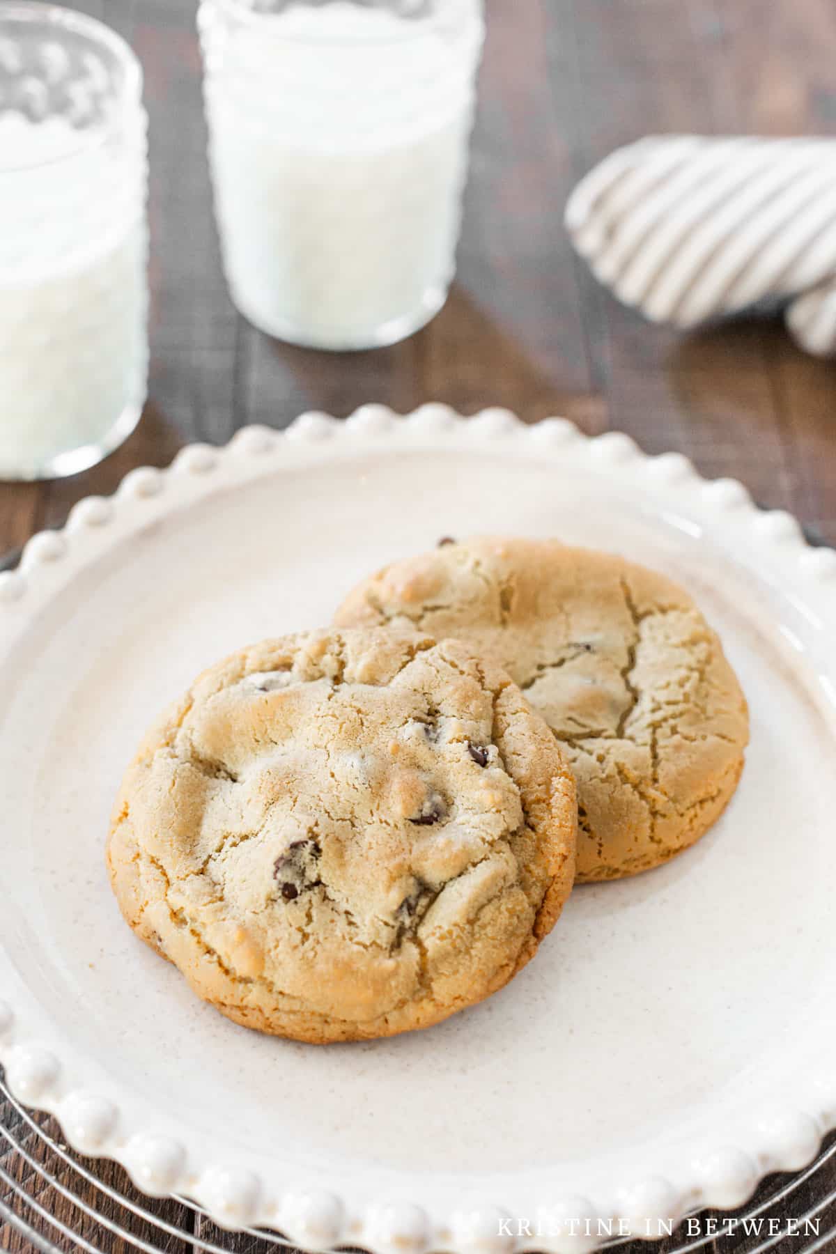 Two cookies sitting on a white plate with two glasses of milk in the background.