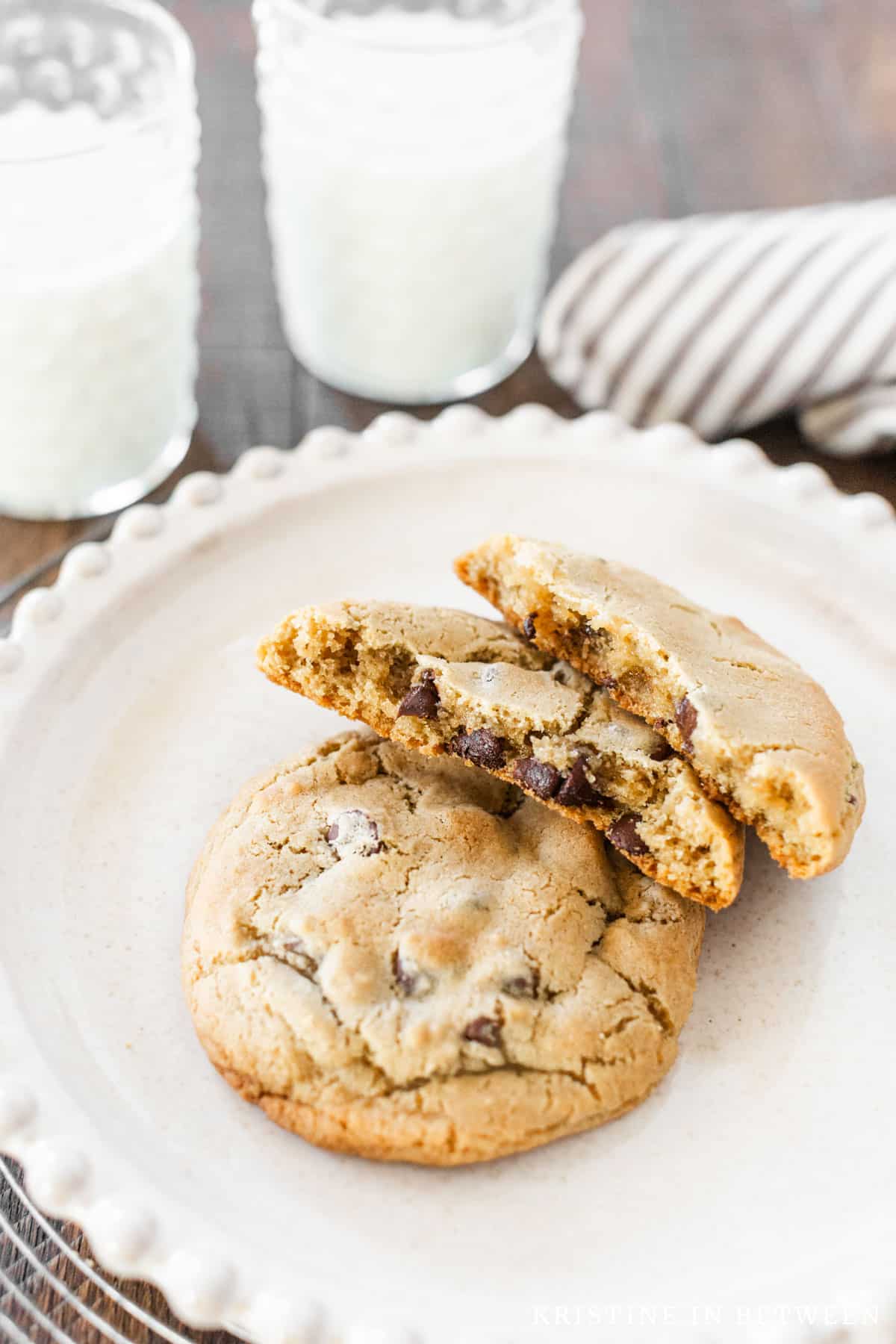 Two chocolate chip cookies sitting on a white plate with one broken in half to show the inside of the cookie.