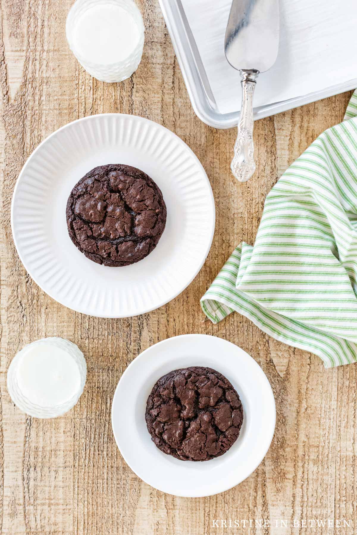 Two dark chocolate cookies sitting on white plates with two glasses of milk and a green striped napkin.