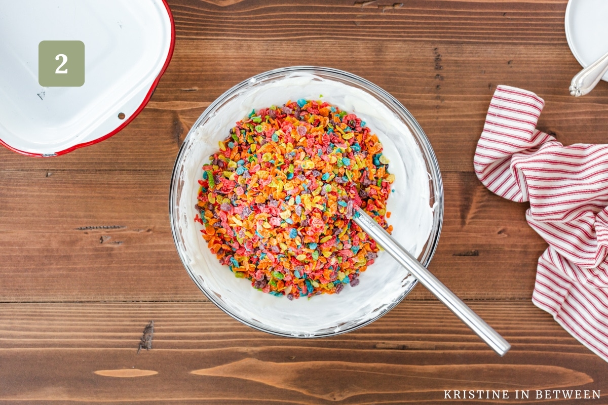 Fruity Pebbles cereal added to the melted marshmallow mixture in a glass bowl.