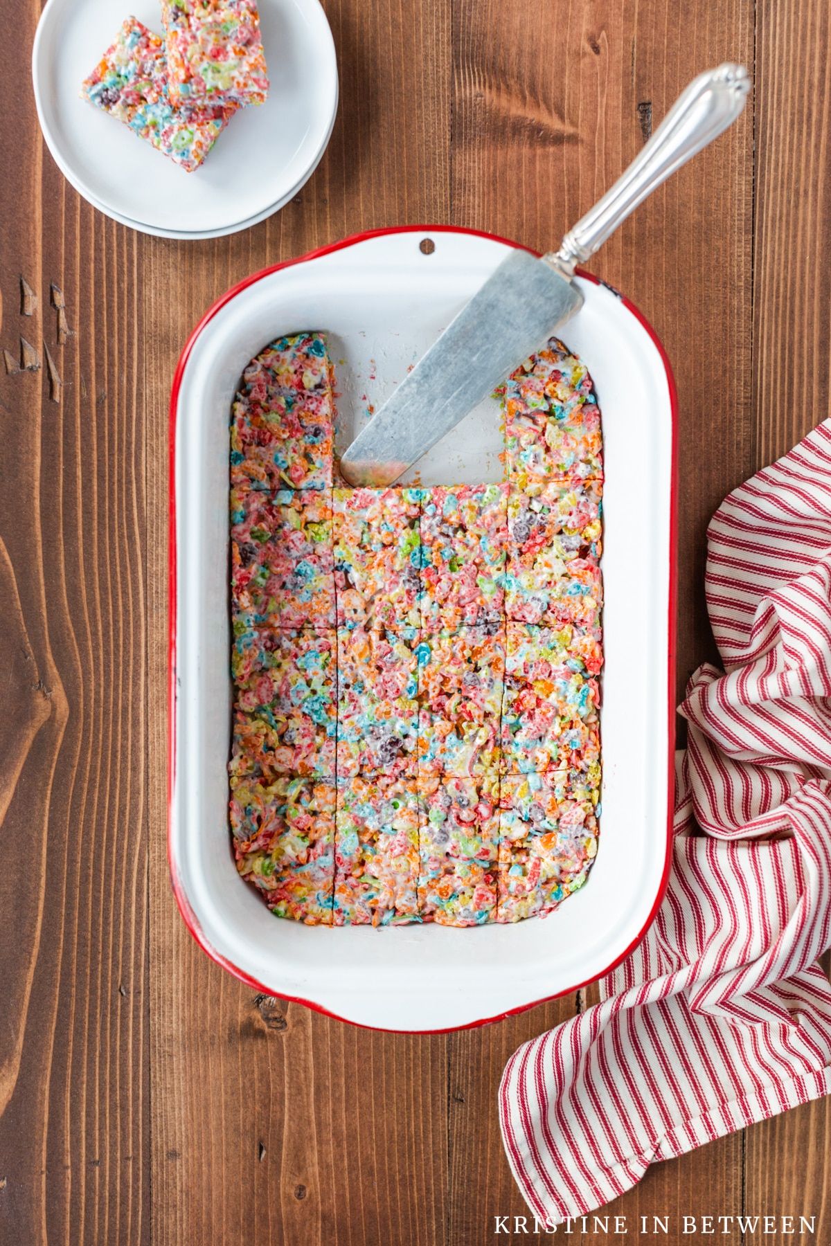 A pan of Fruity Pebbles Krispie Treats with a spatula and a plate of two bars in the background.