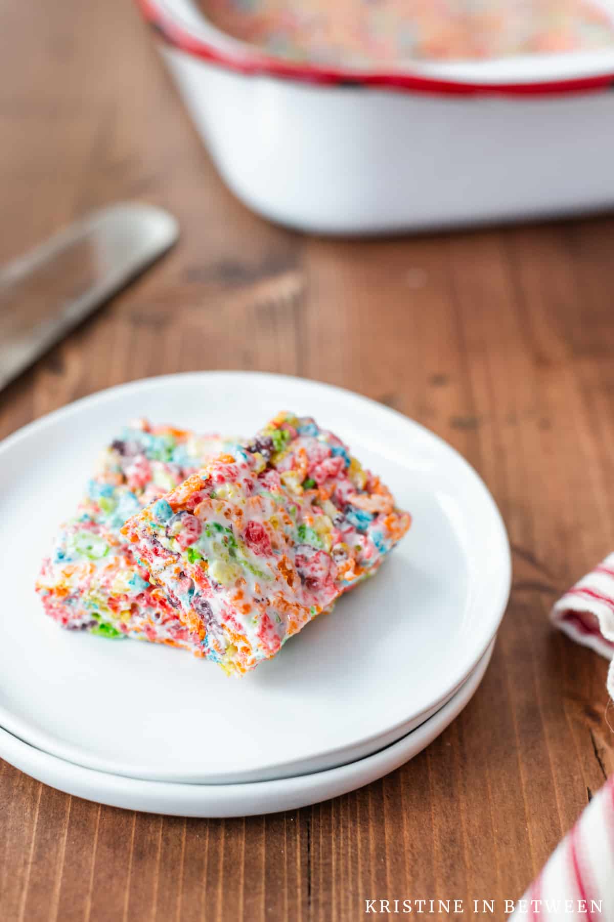 Two Fruity Pebbles Krispie Treats sitting on a white plate with a pan of bars in the background.