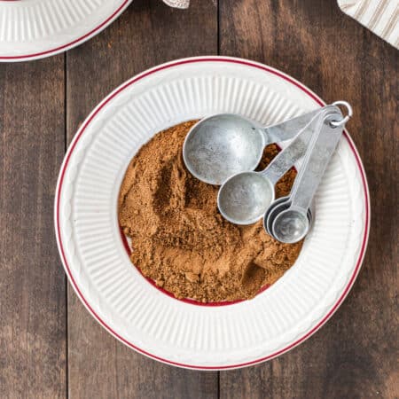 Homemade apple pie spice on a white plate with maroon trim and a small bowl of apples with a spoon in the background.