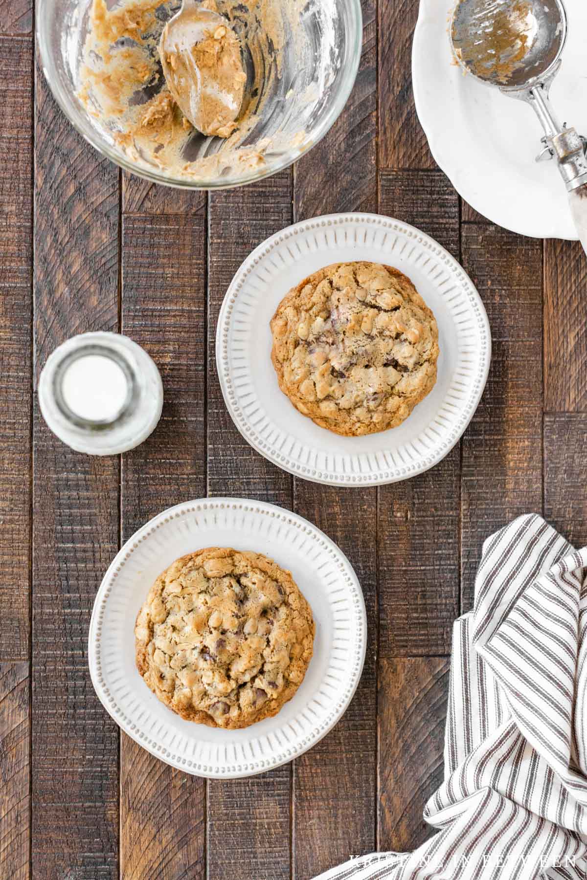 Two large oatmeal cookies sitting on little plates with a bowl in the background.
