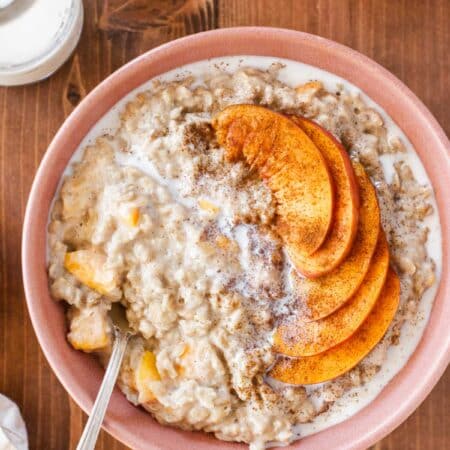 A bowl of peaches and cream oatmeal drizzled with milk and a spoon in it.