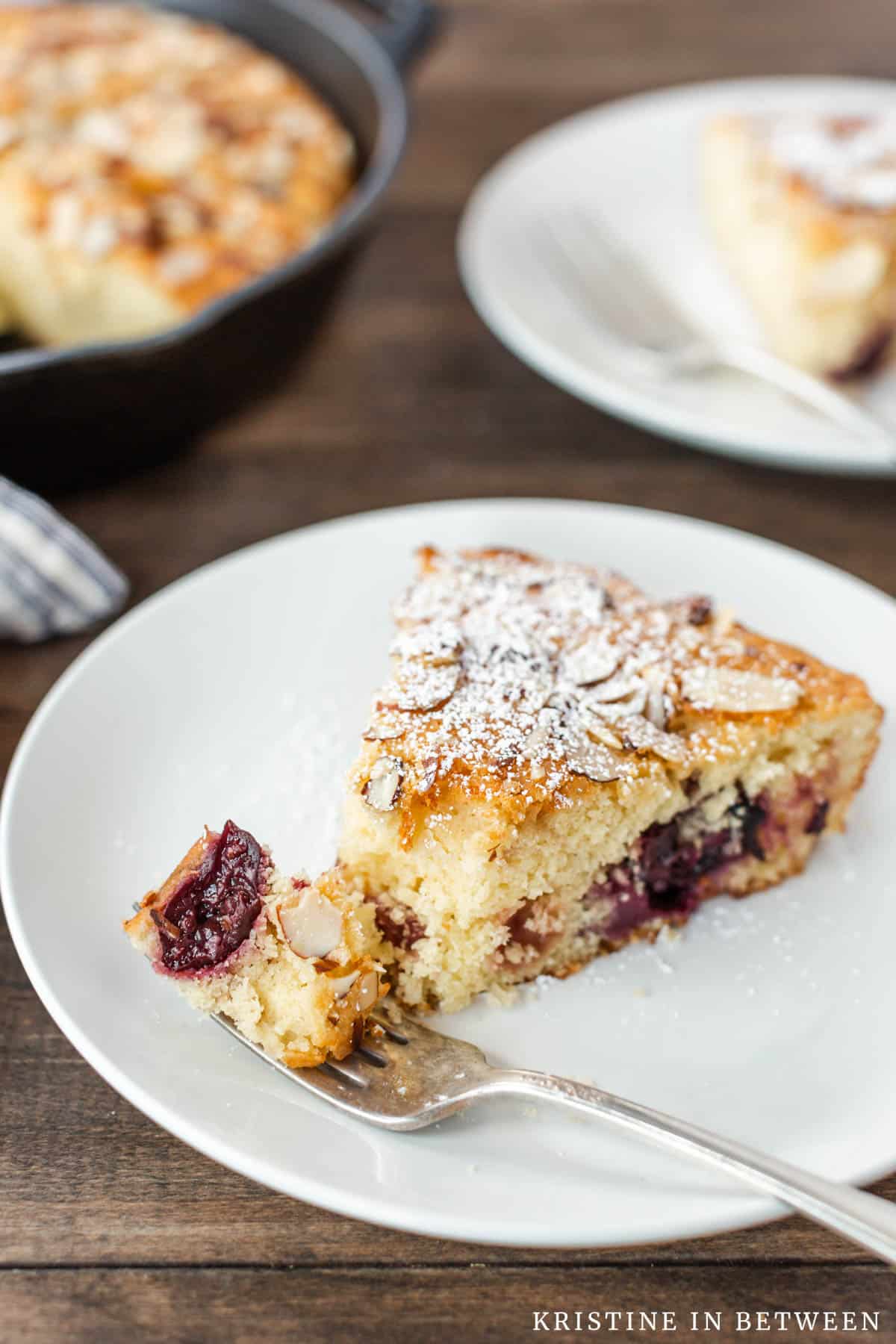 A piece of cherry skillet cake on a white plate with a bite on an antique fork.