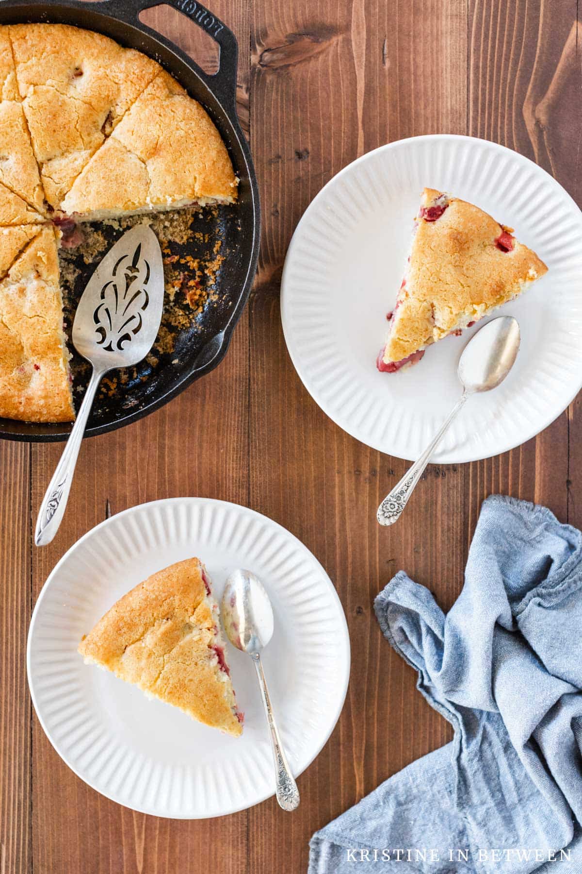 Two slices of skillet cake on white plates with spoons and the rest of the cake in a cast iron pan next to them.