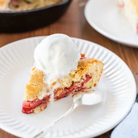 A slice of strawberry skillet cake on a white plate, topped with vanilla ice cream.