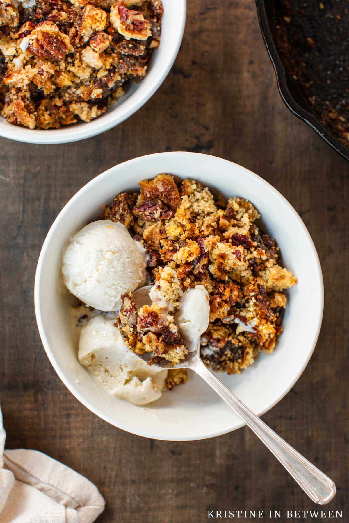 Pecan pie dump cake in a white bowl with two scoops of ice cream and a spoon.