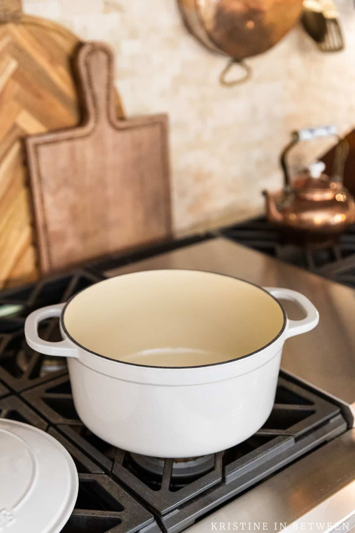 An oyster colored Lodge Dutch oven with the lid off sitting on gas cook top with a copper tea kettle and two cutting boards in the background.
