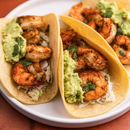 Three air fryer shrimp tacos with avocado on top sitting on a white plate.