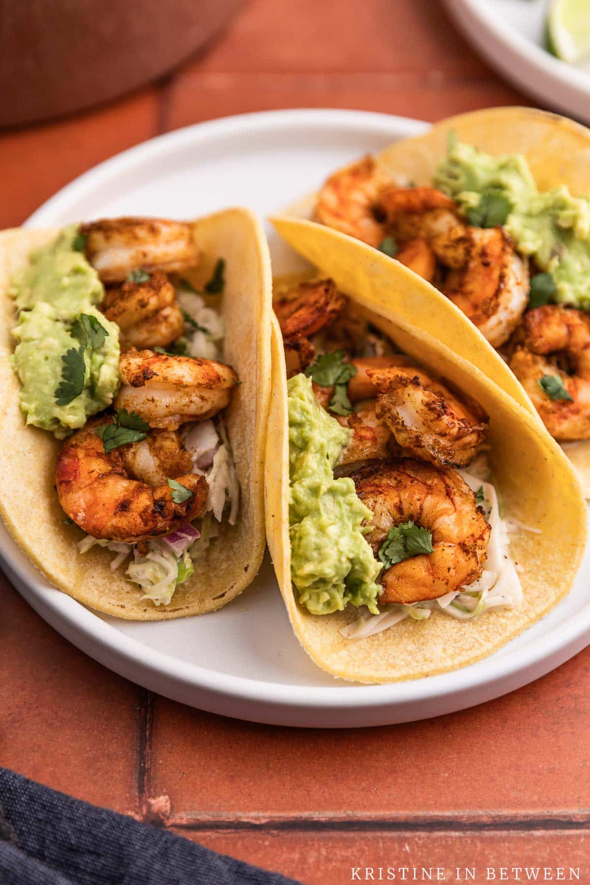 Three shrimp tacos with mashed avocado on top sitting on a white plate.