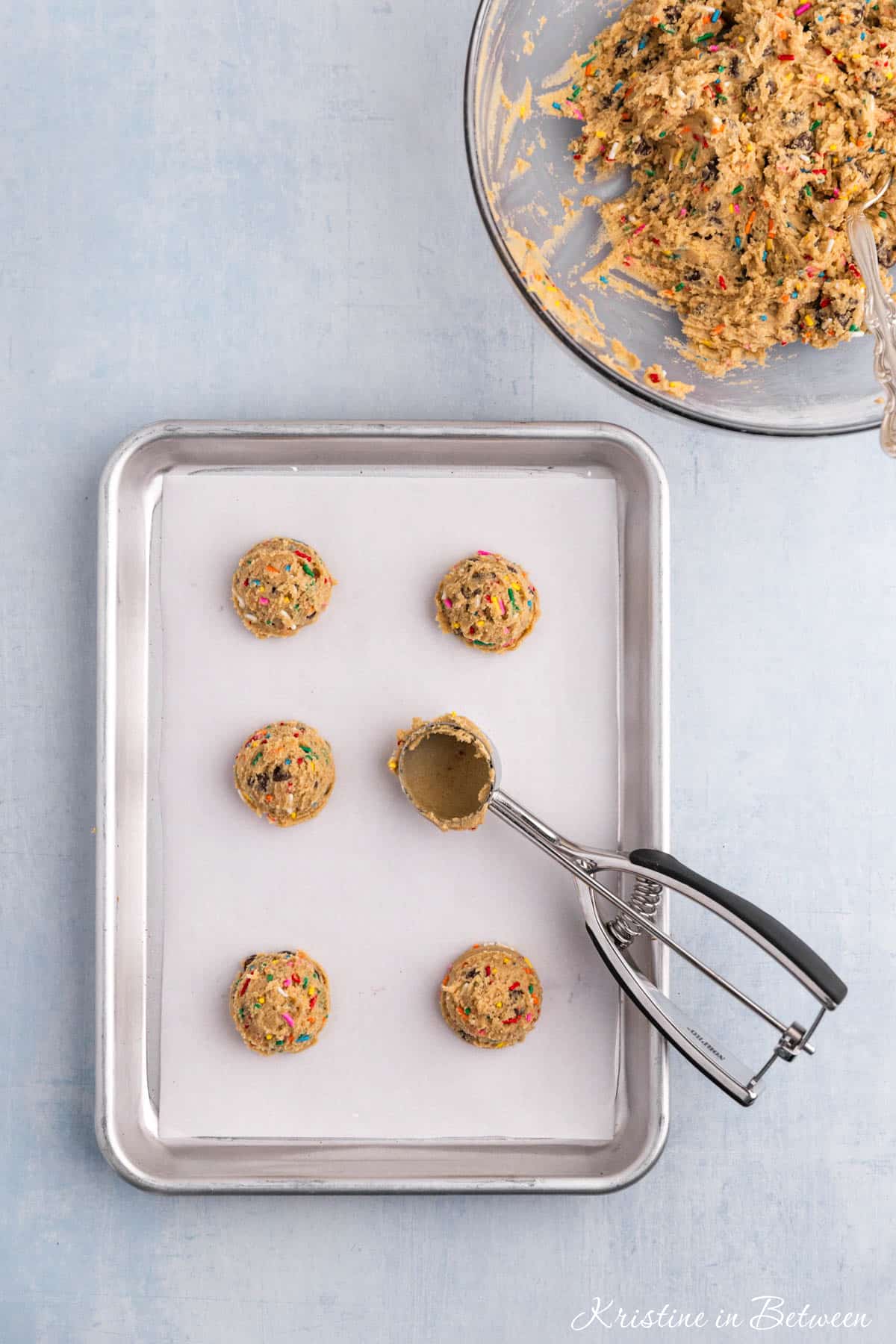Scoops of cookie dough with sprinkles laying on a lined baking sheet with a cookie scoop.