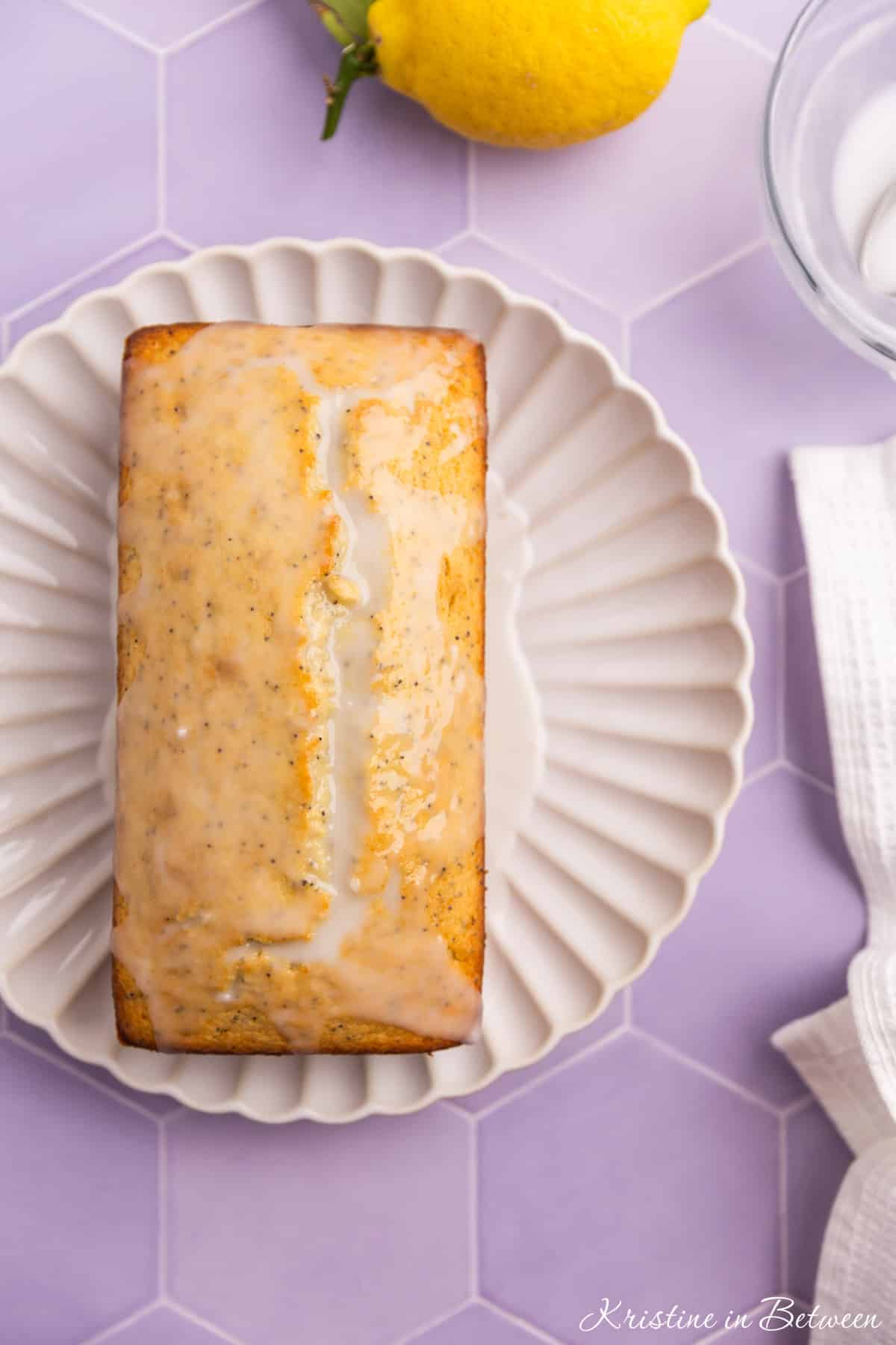 A lemon loaf on a white plate with glaze on top and a lemon in the background.