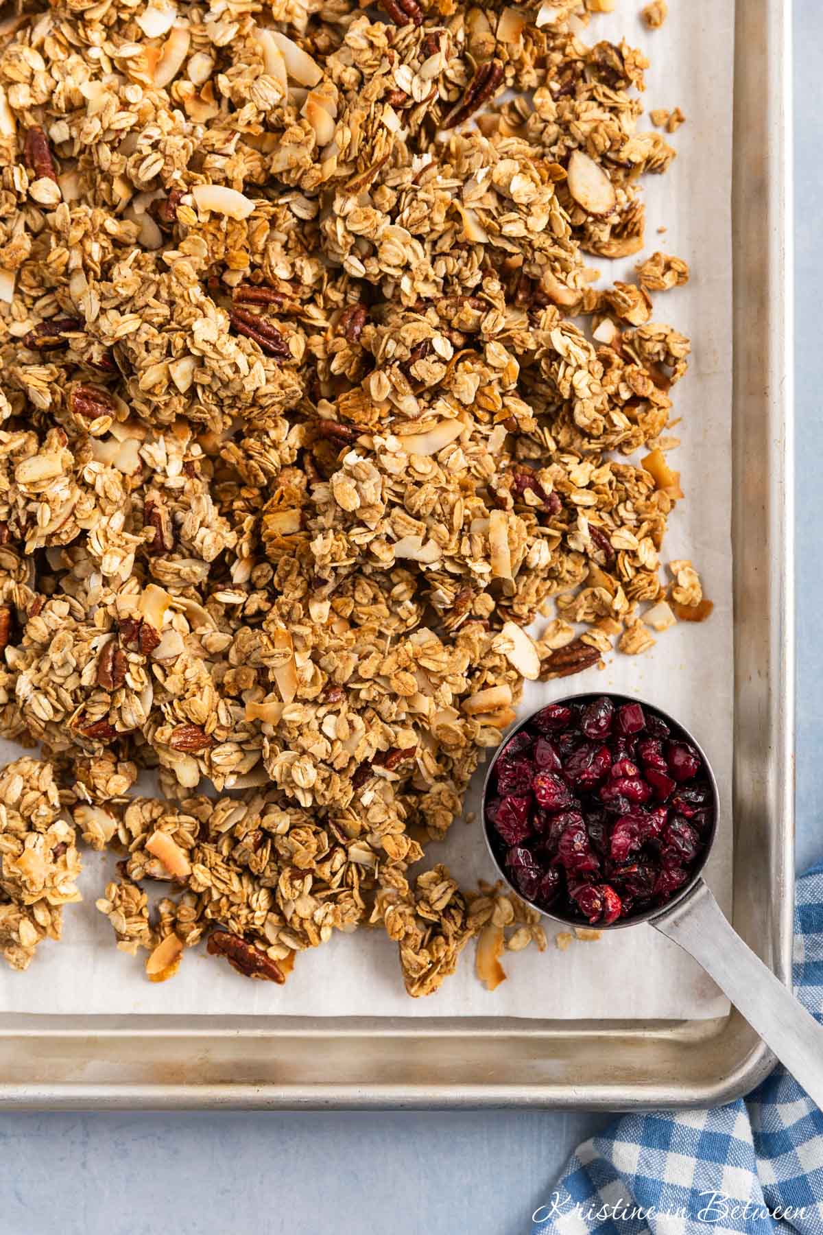 A pan of freshly baked homemade granola on a baking sheet with a small cup of dried cranberries next to it.