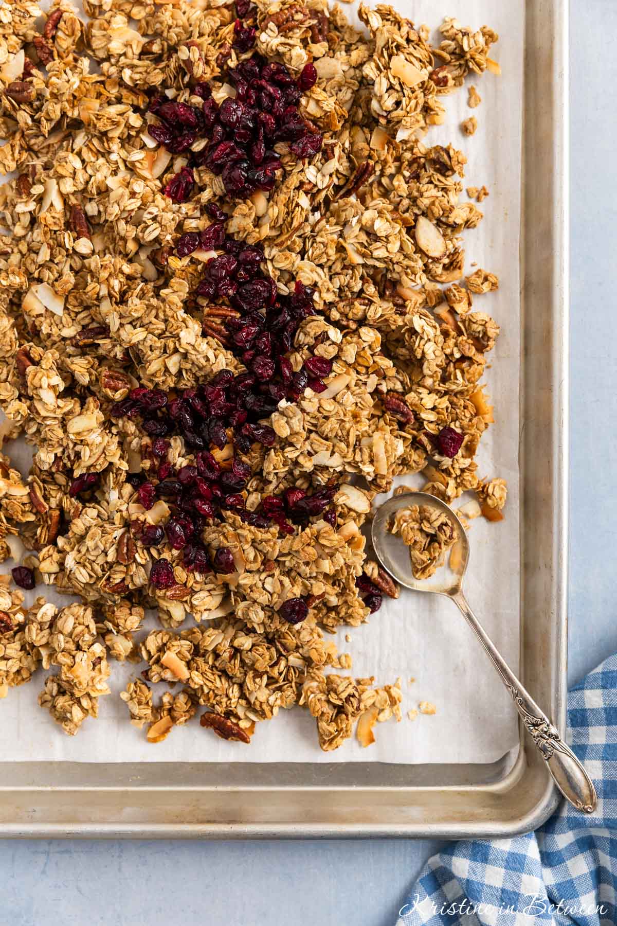 Granola on a baking sheet with dried cranberries on top and a spoon sitting with it.