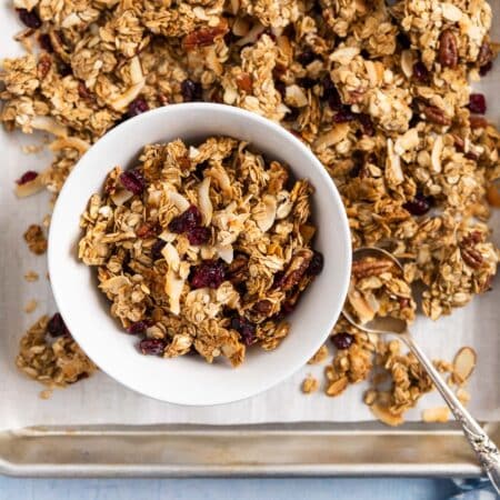 A bowl of freshly baked homemade granola sitting on a baking tray of granola with an antique spoon.