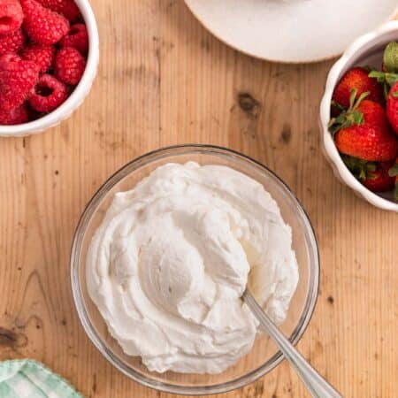 A bowl of homemade whipped cream in a glass bowl with a spoon sitting next to small bowls of berries.