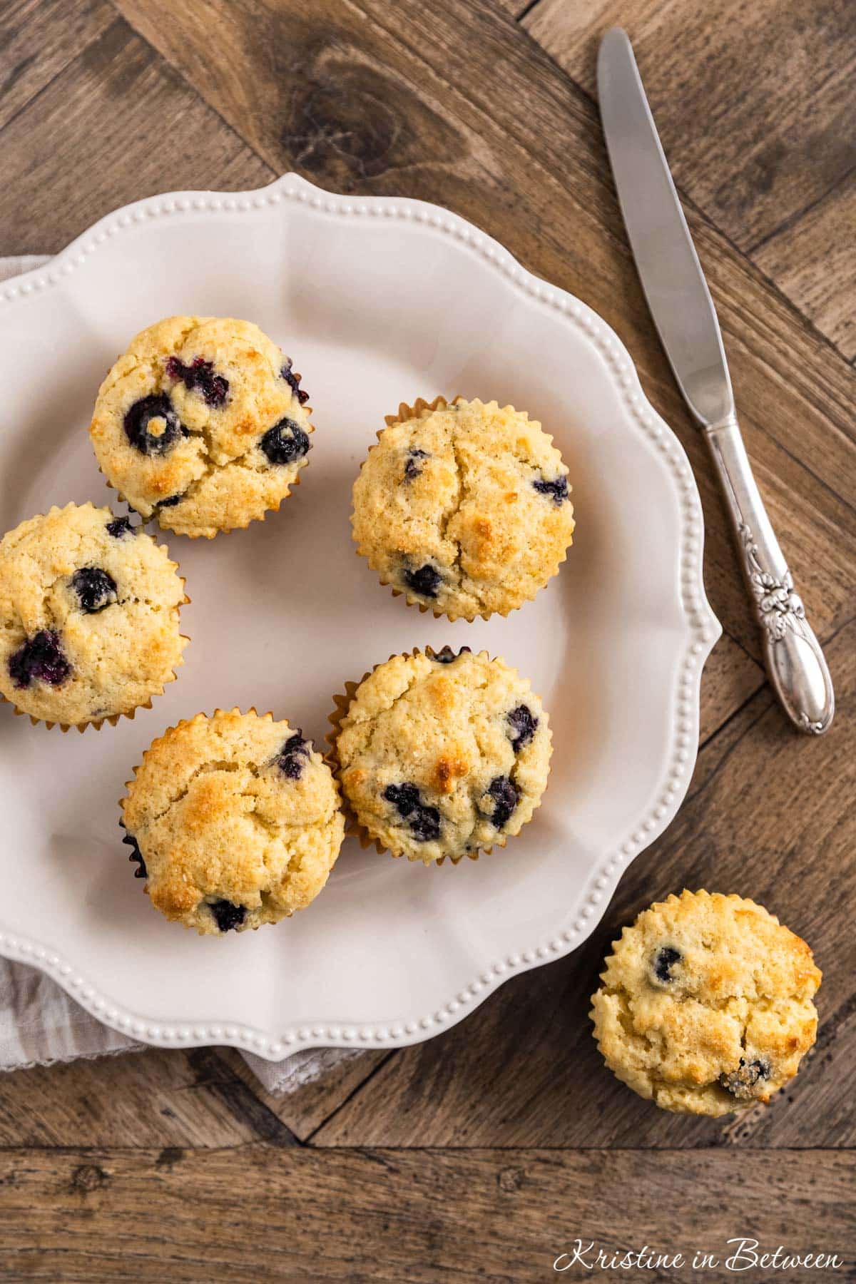 A plate of blueberry lemon muffins with one sitting on the side and a knife in the background.