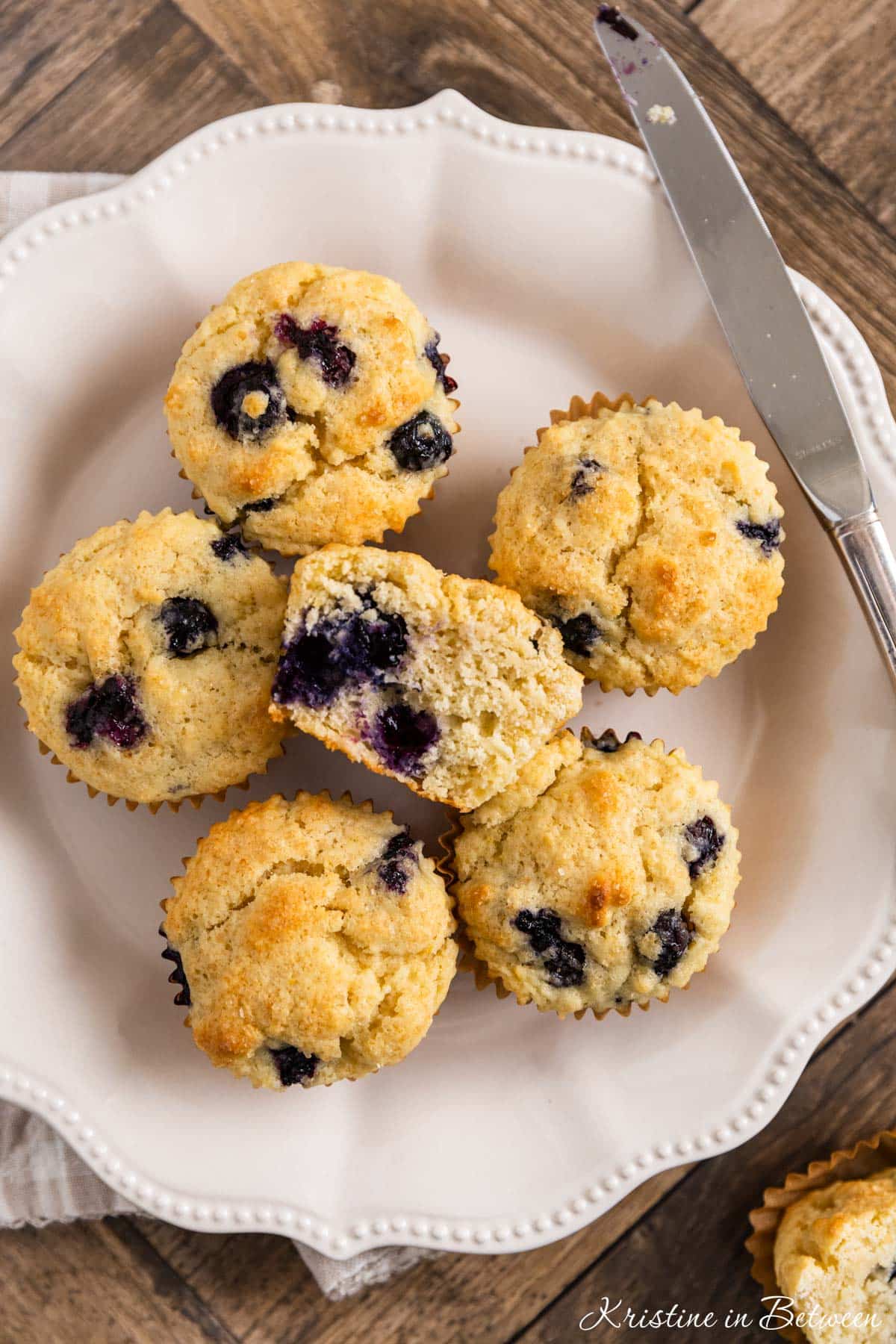 A plate of blueberry lemon muffins with one sitting on the side and a knife in the background.